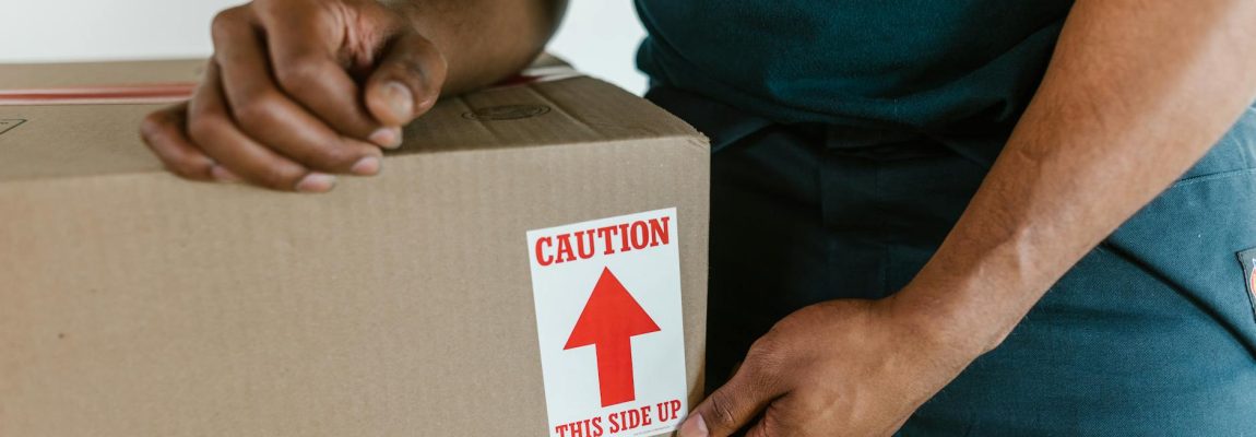 Two professional movers in safety vests carefully pack boxes in a bright, furnished room.