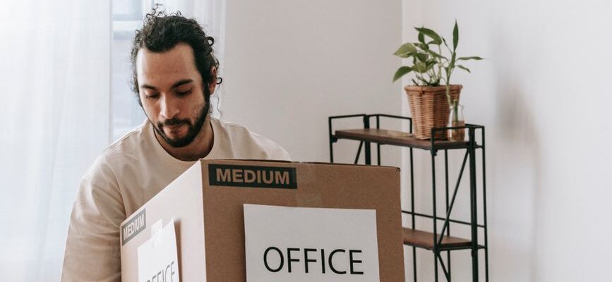 Professional movers carrying furniture into a modern office, with moving truck in background and box labeled "Best House Offi